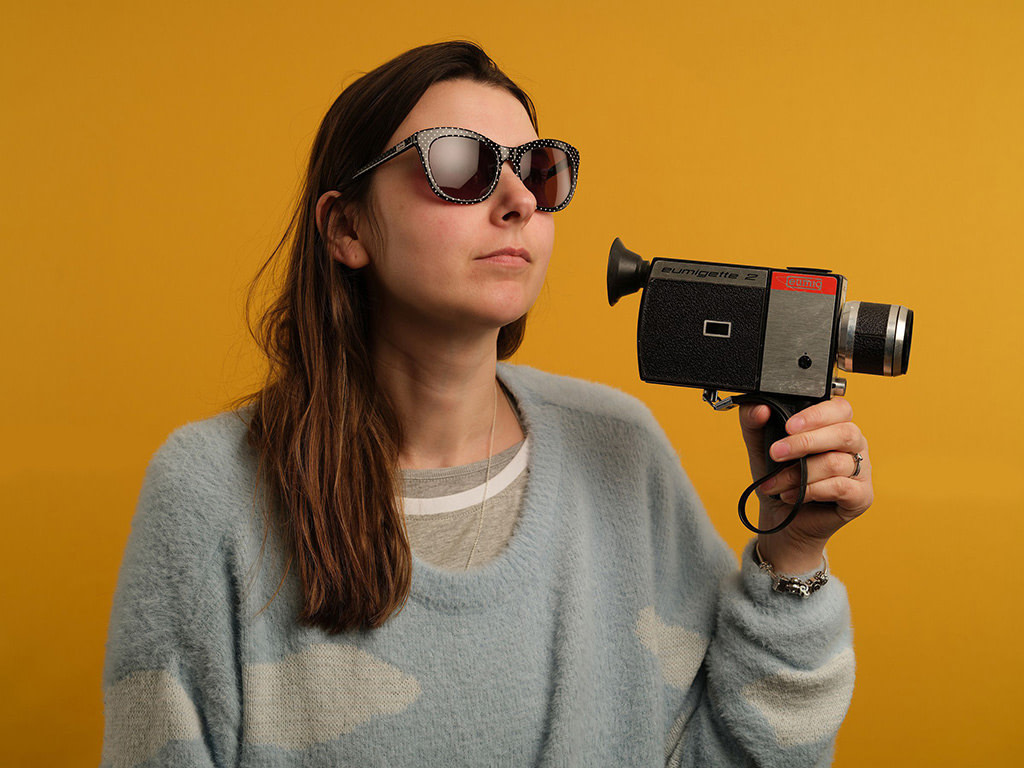 Bright yellow portrait photograph featuring a brunette woman in sunglasses holding a Super-8 camera