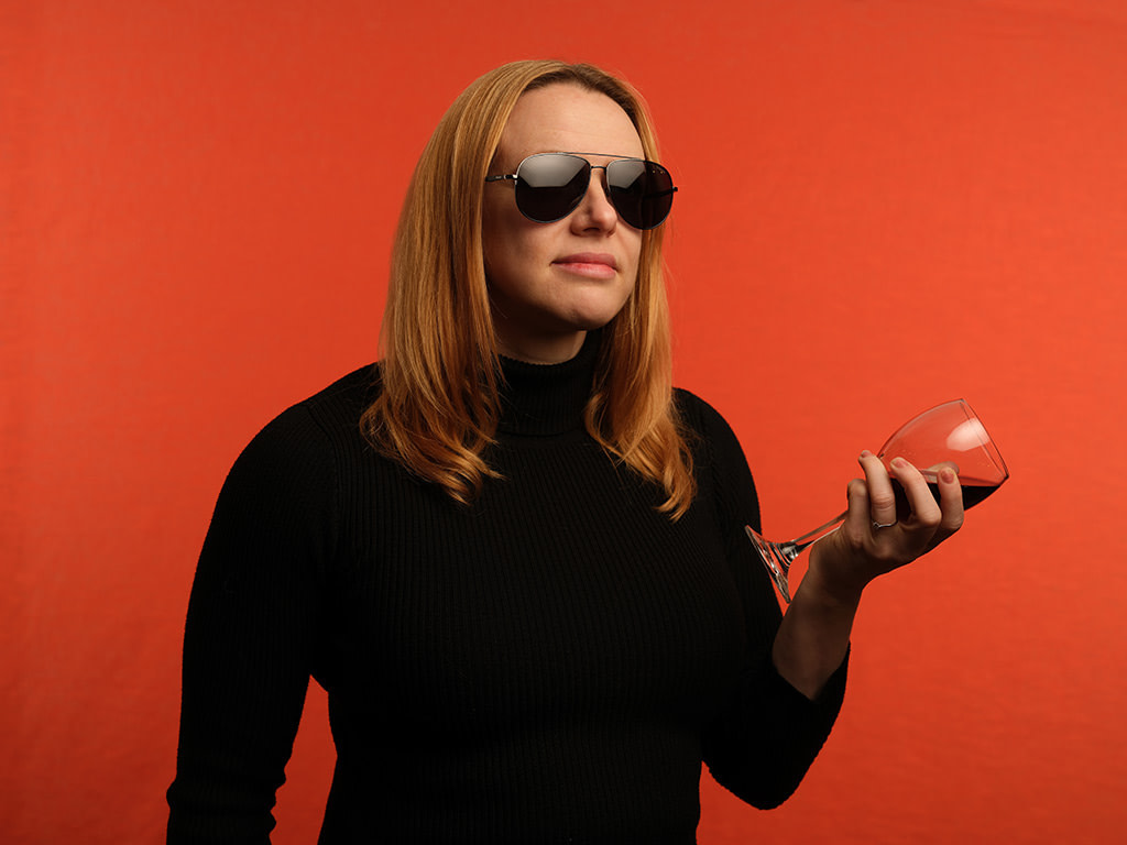 Blond haired woman in sunglasses poses for a studio portrait photograph holding a glass of red wine in front of a vivid orange background