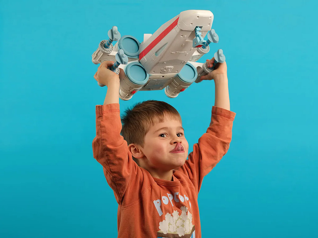 Characterful studio portrait photograph portraying a young boy with his tongue out holding an X-Wing fighter toy above his head in front of a light blue coloured background